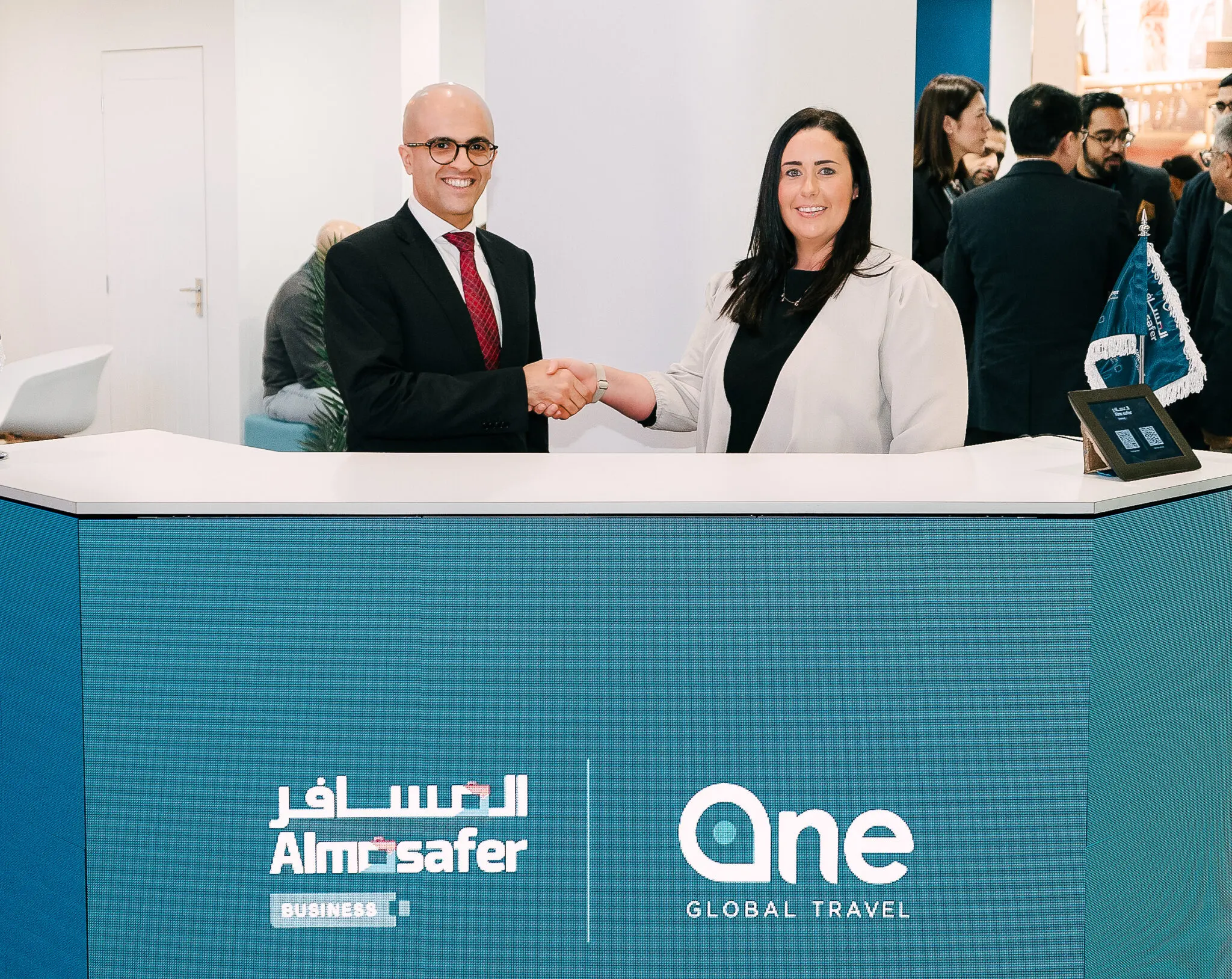 Man and woman shake hands by counter with Almosafer and One Global community logos.