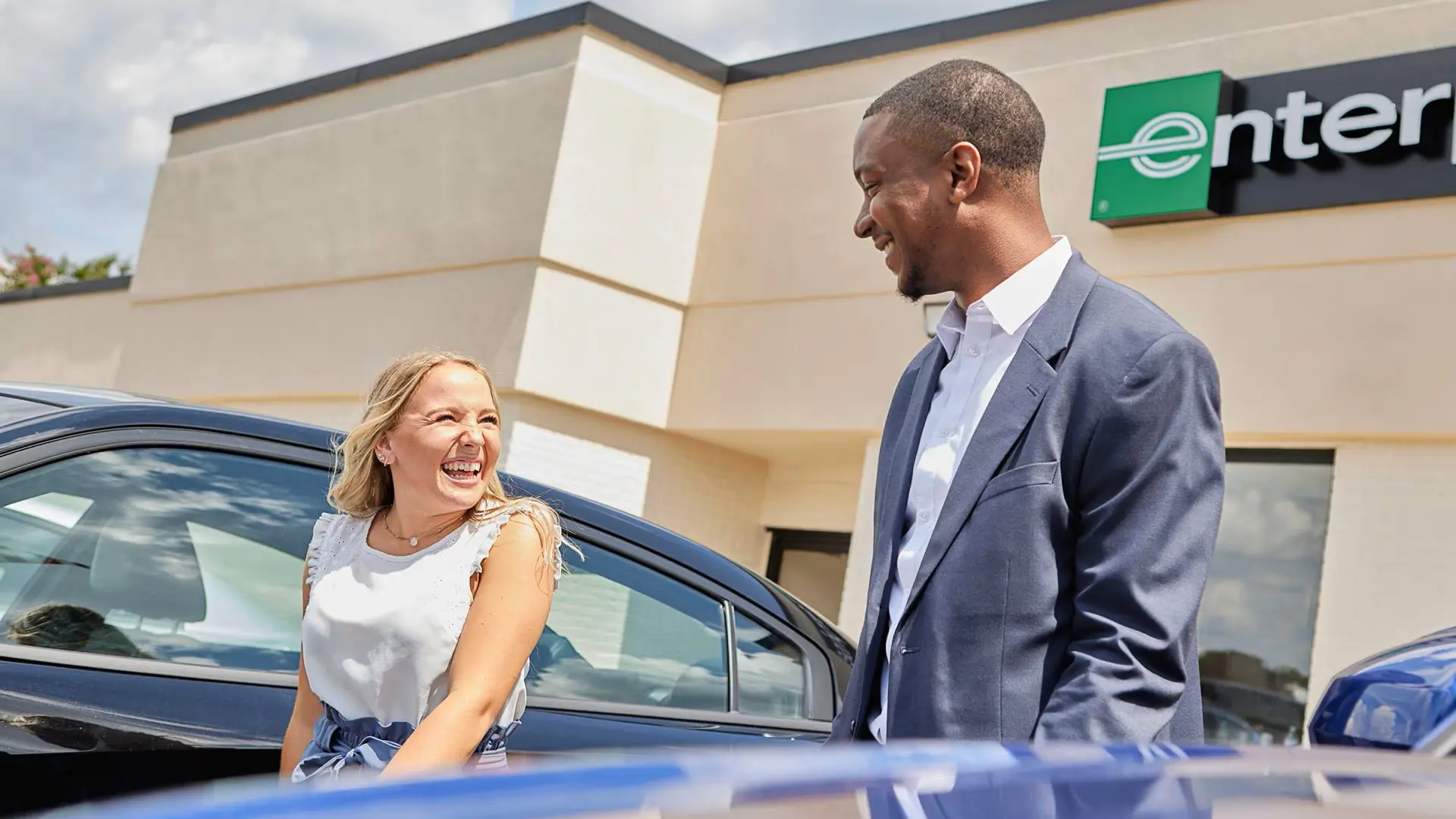 Two people smiling outside a hire location with cars parked in front.