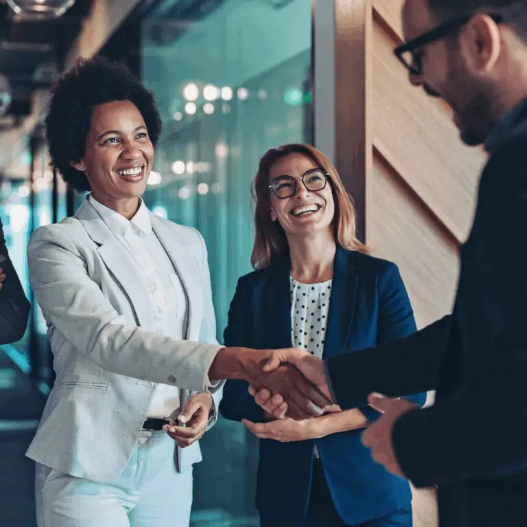 Three professionals smiling and shaking hands in a modern office corridor.