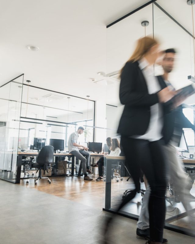 Two people walk through One Global Way office with glass walls and busy desks.