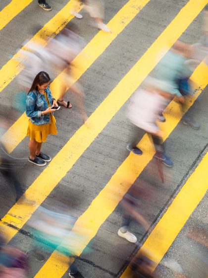 A woman, stationary on a crosswalk as people move past her, looking down to her smartphone.